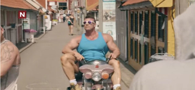 A huge Norwegian body builder on a tiny bike in a tiny town with tiny buildings. 